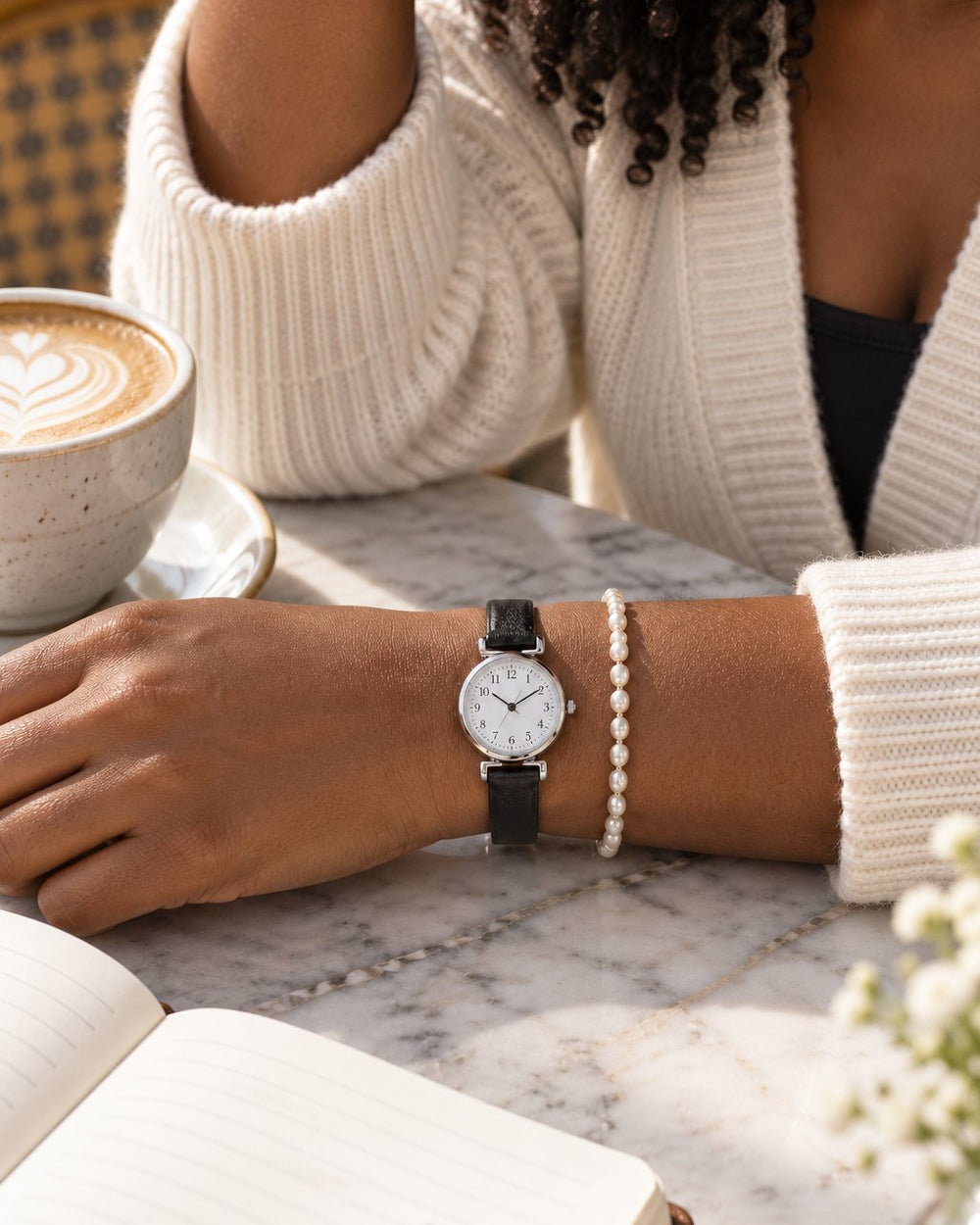 Wrist with the petite watch and a pearl-look bracelet resting on a café table beside a latte