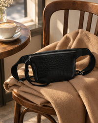 Black croc-embossed belt bag resting on a folded camel coat beside a coffee cup