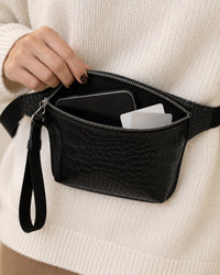 A hand holds the slim belt bag open with a phone and cards tucked inside