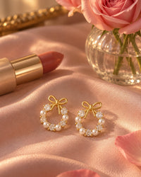The bow wreath studs on a blush satin cloth beside a lipstick and a small vase of roses