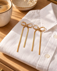 The gold-tone chain bow earrings on a folded white shirt beside a ceramic dish and a coffee cup