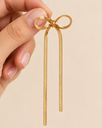 One chain bow earring held up between two fingers, its long tails hanging down