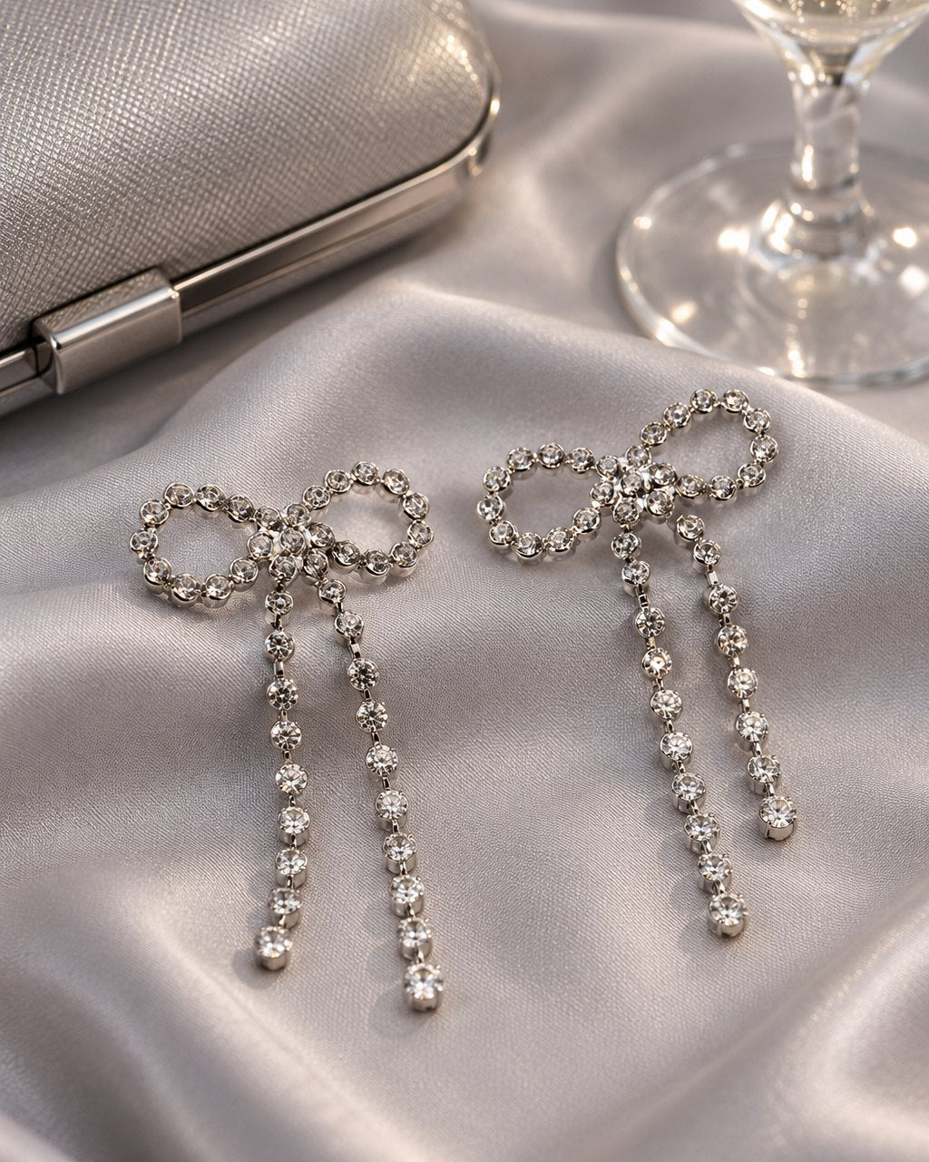 The silver-tone sparkle bow earrings on a pale grey satin cloth beside a small evening clutch and a glass of bubbly