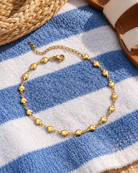 The heart anklet on a striped beach towel beside a straw tote and flat sandals