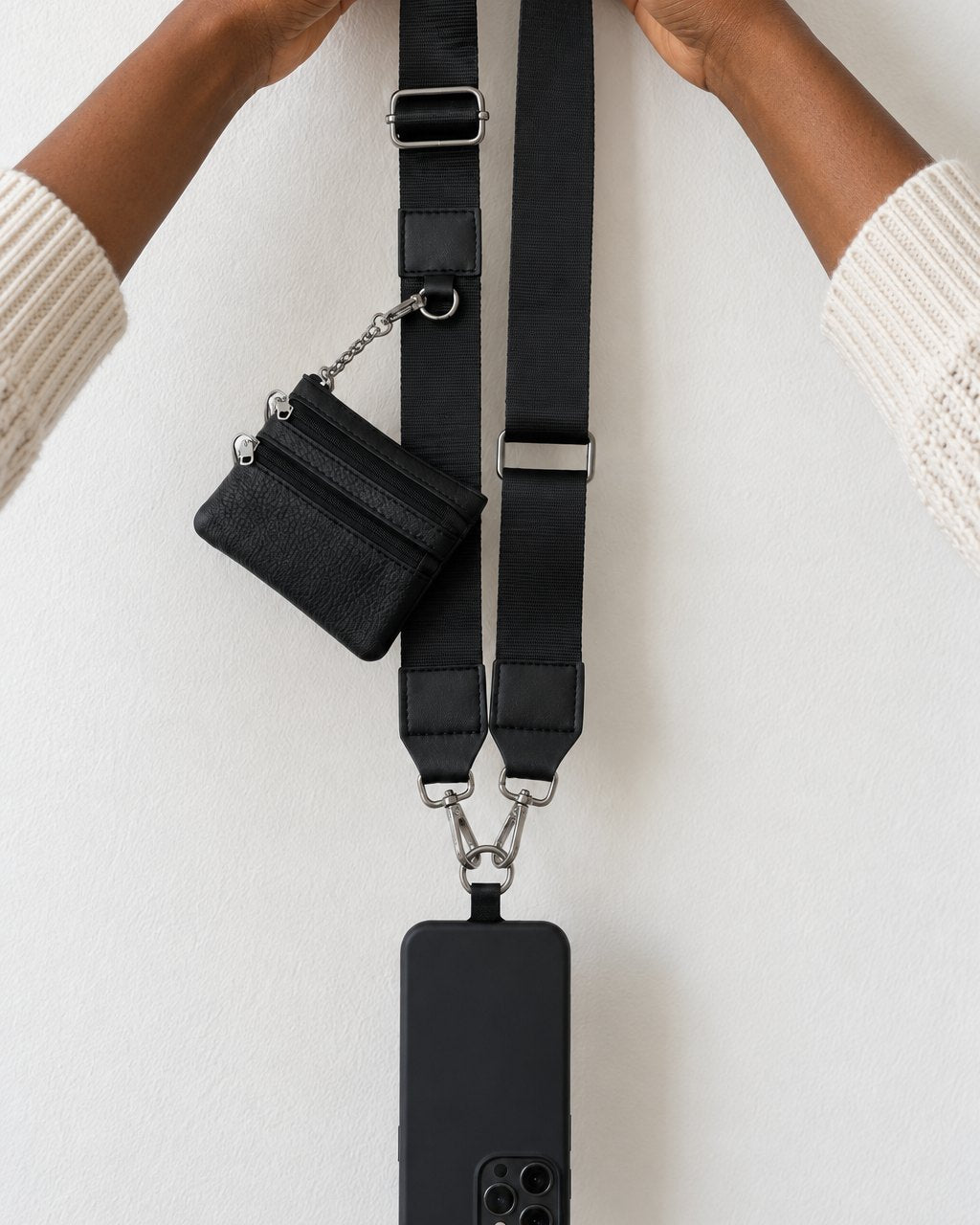 Hands holding up the black strap with a phone hanging from it and the small zip pouch clipped on