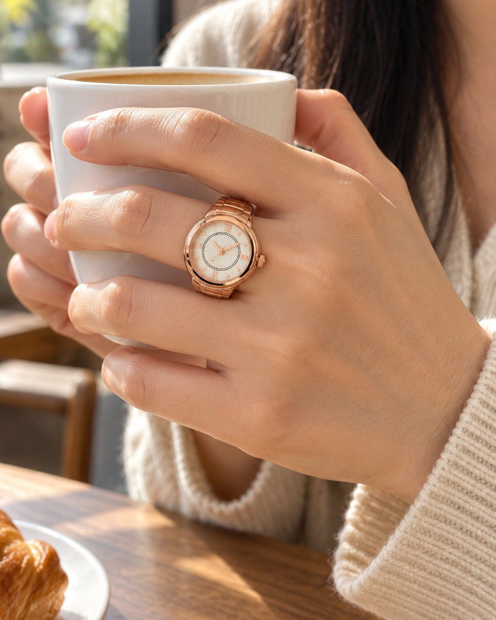 A hand holding a coffee cup tilts the ring watch on her middle finger toward the light