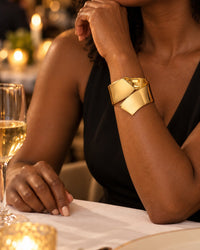 Woman in a black sleeveless dress at a restaurant table, wearing the gold-tone cuff alone on her wrist