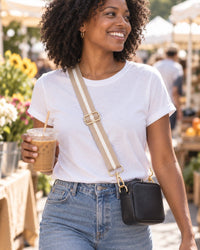 Woman at a market wearing a small bag crossbody on the beige striped strap, coffee in hand